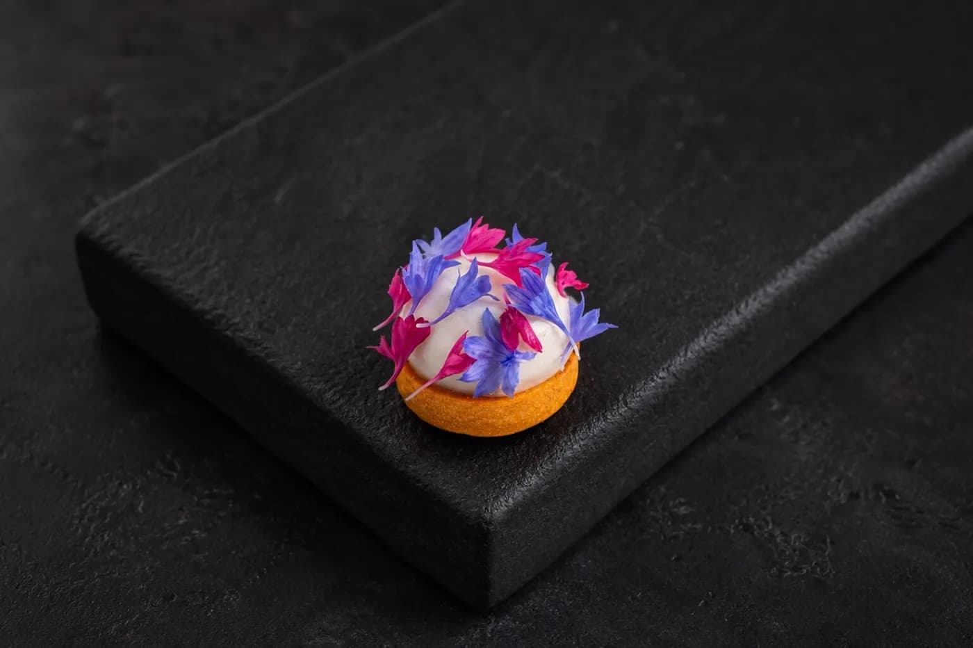 A tartlet dressed in blue and magenta petals on black stone